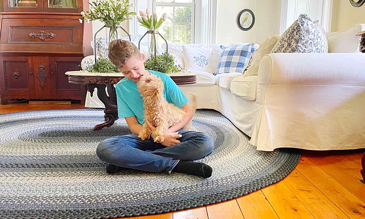 A boy is sitting on a braided wool rug in the living room with his dog.