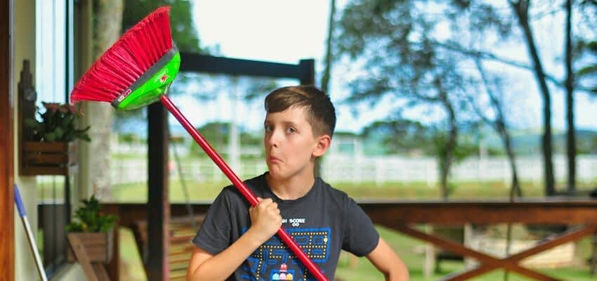 A child is holding a broom on a front porch, feeling silly after doing some floor sweeping. He is making a fun expression on his face.