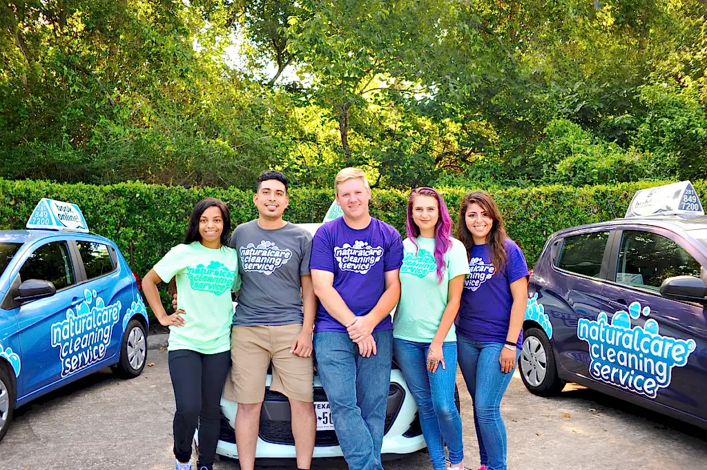 A group of maids from Naturalcare Cleaning Service are posing for a photo in front of their branded vehicles, in a parking lot on a summer day. They are cleaners Rosenberg TX sees often.