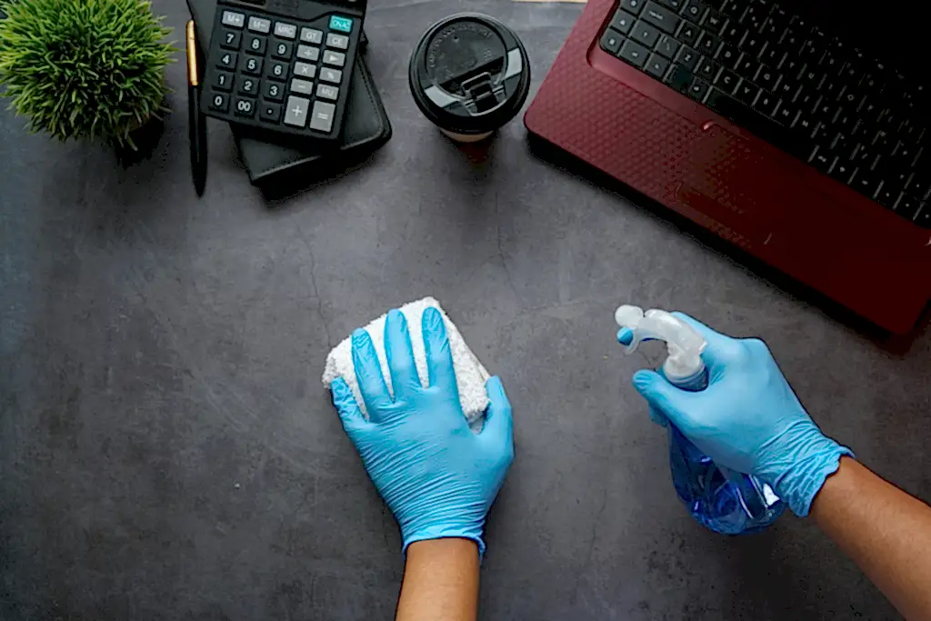 Hands adorned in latex gloves are disinfecting and scrubbing an office desk. These gloves are recommended to use in office cleaning safety tips.