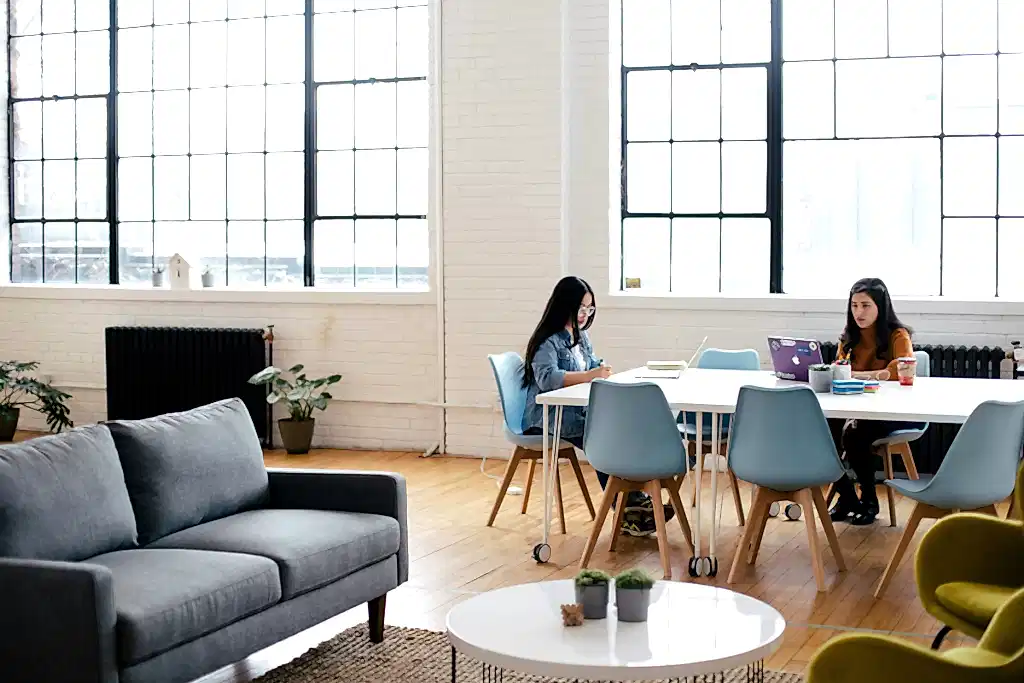 Two young professionals work on laptops in a coworking space. They're benefitting from using office cleaning tips to improve their workspace.