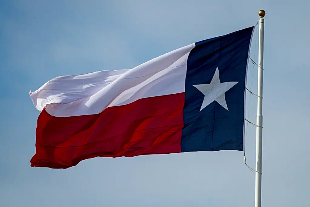 A Texas flag is flying in the wind. A blue sky is behind it.